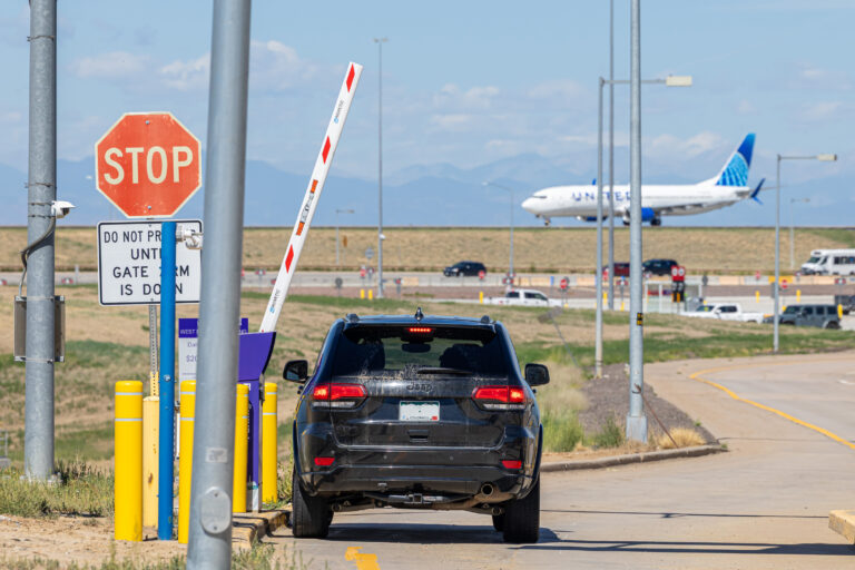 A black SUV waits at a stop sign near an airport gate as a United Airlines plane takes off in the background.